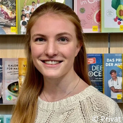 Eine lächelnde junge Frau mit langen braunen Haaren steht vor einem Bücherregal voller verschiedener Bücher. Sie trägt ein entspanntes Outfit und blickt direkt in die Kamera. Ihr Name ist Katharina Kahl.